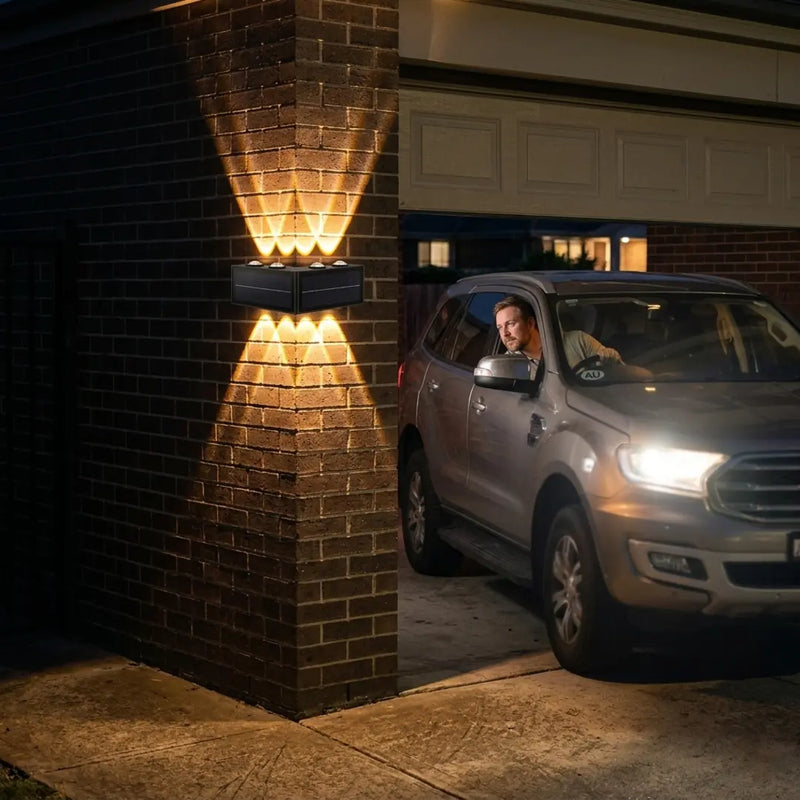 A man driving a silver SUV into a garage, with the PT0803 Grivo solar corner light mounted on the dark brick entrance casting warm up and down beams to illuminate the pathway, demonstrating its functionality as a safety and ambient garage light.