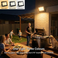 E027EI Voro floodlight mounted on a brick wall above an outdoor BBQ, glowing warm 3000K amber, family and friends gathered on a timber deck at night with fairy lights overhead, inset shows all three colour options: 3000K Warm, 4000K Natural, 6000K Bright