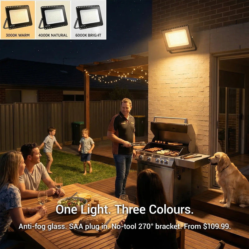 E027EI Voro floodlight mounted on a brick wall above an outdoor BBQ, glowing warm 3000K amber, family and friends gathered on a timber deck at night with fairy lights overhead, inset shows all three colour options: 3000K Warm, 4000K Natural, 6000K Bright