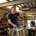 A warehouse worker is installing an Outway high bay LED light from a ladder inside a storage facility.