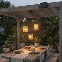 Three woven pendant lights hang over a wooden dining table under a pergola in a garden setting, with a small solar panel mounted on the pergola beam above. The lights cast a warm glow, illuminating the table setting and surrounding plants at dusk.