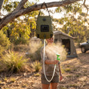 Camper smiles while spraying mist from solar shower bag; sunlight filters through leaves, highlighting portable outdoor hygiene setup.