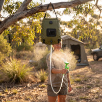 Camper smiles while spraying mist from solar shower bag; sunlight filters through leaves, highlighting portable outdoor hygiene setup.