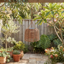 Solar-powered woven rattan hanging lantern with visible solar panel on top, suspended under a wooden pergola in a sunlit garden with tropical plants and wooden fence background.
