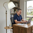 FL068 antique brass floor lamp with wood side tray positioned beside a timber desk, cool directed light, man working on laptop in a home office with bookshelves