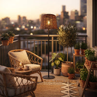 Stylish apartment balcony setup featuring the PT0908 Solar Floor Lamp, rattan armchair, and potted olive tree against a city skyline at sunset. Highlights the lamp's boho aesthetic and suitability for urban outdoor living areas without wiring.