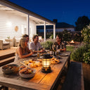Friends gather around outdoor dining table at night, lit by solar lantern; fire pit glows softly in garden background.