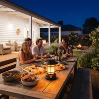 Friends gather around outdoor dining table at night, lit by solar lantern; fire pit glows softly in garden background.