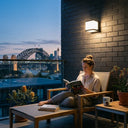 Woman reading on a city apartment balcony at night with a PT0711 Cube solar wall light illuminating the wall, Sydney Harbour Bridge visible in the background.