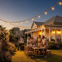 Friends gather for BBQ under solar string lights strung above lawn; white cottage glows warmly in background as dusk settles.