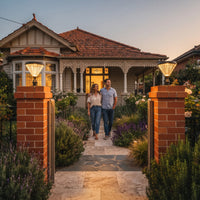 A couple walks along a stone path toward a Victorian-style house, framed by two brick pillars with glowing outdoor lights and landscaped garden beds at dusk.