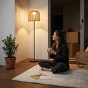 A woman sitting cross-legged on an indoor rug holding a coffee cup, with the PT0908 Floor Lamp beside her casting beautiful rattan shadows, highlighting its warmth and artistic value as an indoor decor piece.