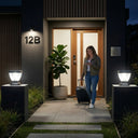 Airbnb guest with rolling suitcase checking phone at front door of modern home numbered 12B at night, two Nexi Solar Pillar Lights PT0112 glowing on dark rendered pillars flanking entrance path