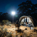 Campers relax near tent lit by SolarFlex Pro on tripod; starry sky and bushland surround the glowing campsite scene.