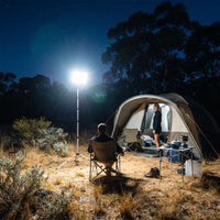 Campers relax near tent lit by SolarFlex Pro on tripod; starry sky and bushland surround the glowing campsite scene.