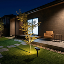 A modern outdoor patio scene at night with a small tree illuminated by a ground-mounted solar landscape light. The setting features a dark wood-paneled house, a stone path, a brown leather armchair, and a side table.