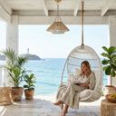 A woman lounges in a white hanging egg chair on a covered porch with a view of the ocean. She is wearing a beige robe and reading a book, with potted plants, a woven pendant light, and a stone floor around her.