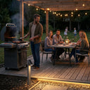 Rivix PT0723 solar bollard light placed on a timber deck beside a BBQ grill, with a group of friends dining under string lights on a pergola while children play on the lawn in the background.