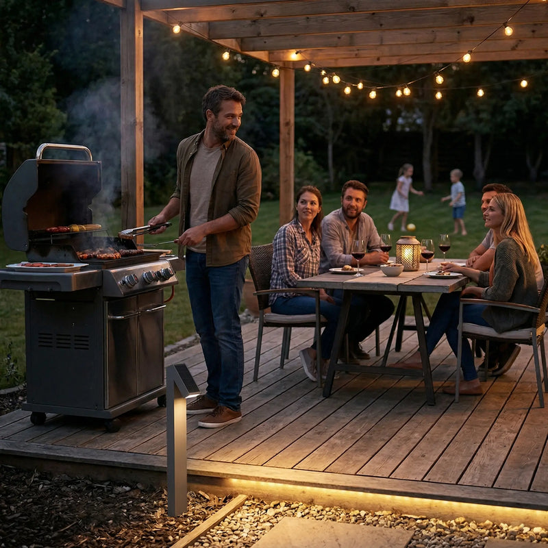 Rivix PT0723 solar bollard light placed on a timber deck beside a BBQ grill, with a group of friends dining under string lights on a pergola while children play on the lawn in the background.
