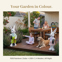 Lumi A002 solar rainbow rabbit garden ornament boy and girl pair on a backyard deck at dusk with a family having dinner a golden retriever and a young girl pointing at the rabbit with text overlay Your Garden in Colour RGB Rainbow Solar USB-C 4 Modes All Night
