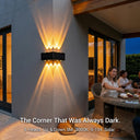 A group of people enjoying an outdoor dinner party, with the PT0803 Grivo solar corner light mounted on a grey wall in the background casting a warm glow. The overlay text 'The Corner That Was Always Dark. 8 Heads. Up & Down 5M. 3000K. 9-11H. Solar.' highlights its ability to eliminate dark spots with long-lasting, smart solar lighting.