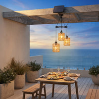 A three-light outdoor pendant chandelier with woven crystal-like shades and a small solar panel on the ceiling, hanging over a dining table on a wooden patio with an ocean view at sunset.