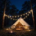 Glamping tent nestled in forest at night, lit by solar string lights strung between trees; campfire flickers nearby under starry sky.