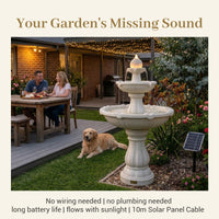 A garden fountain product photo with marketing text. The scene shows a stone-style tiered fountain on a lawn beside a patio, with a couple dining at a table, a dog lying nearby, and a small solar panel.