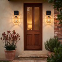 Symmetrical pair of PT0909 solar wall lights flanking wooden front door, casting warm woven shadows on beige stucco walls; potted rosemary and kangaroo paw plants frame entrance — elegant, wire-free curb appeal.