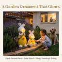 OUTWAY Hoppi A004 solar bubble rabbit ornament pair glowing warm LED light in a lavender garden at dusk with a young girl in pyjamas and a golden retriever with a woman watching from the kitchen window and text overlay A Garden Ornament That Glows Hand-Painted Resin Solar Glow 8-10hrs Standing and Sitting