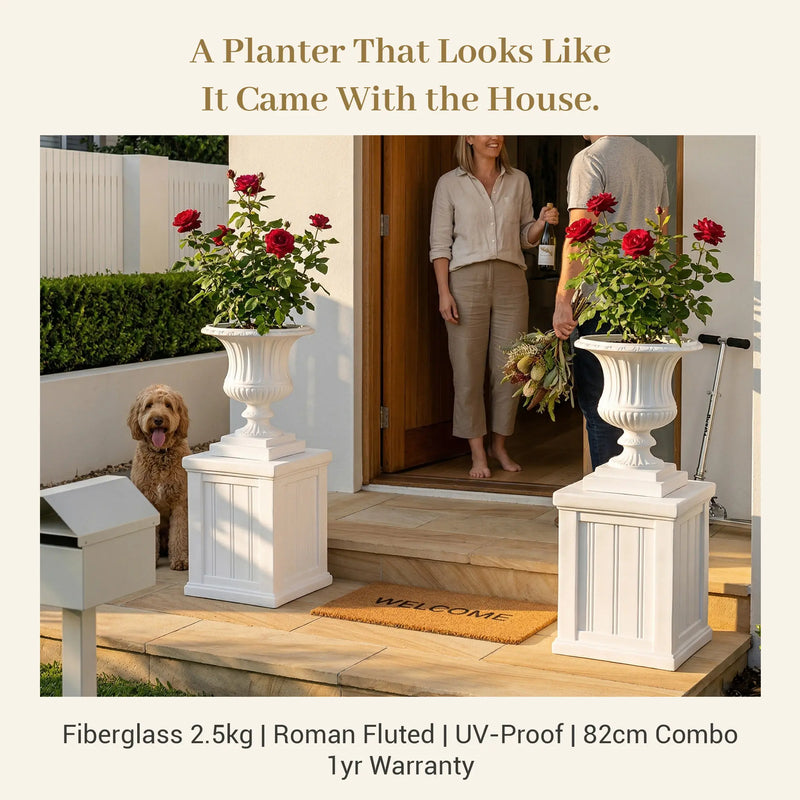 OUTWAY Flora Roman planter combo flanking front door with red roses, couple welcoming guests, headline: "A Planter That Looks Like It Came With the House", fiberglass 2.5kg UV-proof 82cm specs — F004-F006-flora-combo-hero-entrance-couple-roses
