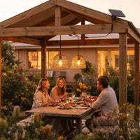 Three friends dining under PT0914 lanterns in wooden gazebo at sunset, wine glasses and floral centerpiece — ideal for intimate gatherings with natural texture and warm glow.