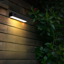 A rectangular black outdoor wall light mounted on a dark wooden wall, casting a warm yellow glow, with green foliage and a red flower in the foreground at night.