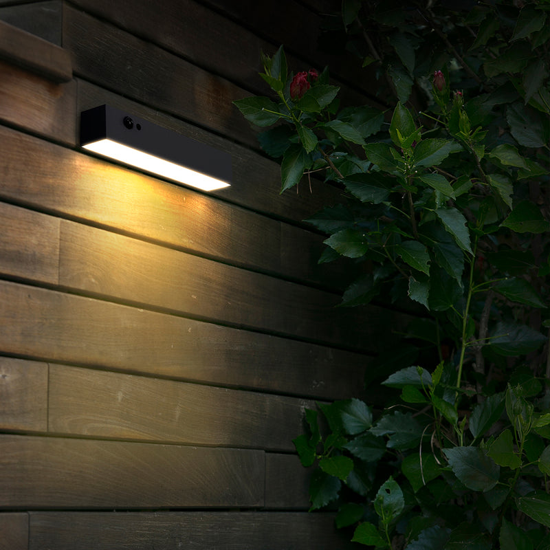 A rectangular black outdoor wall light mounted on a dark wooden wall, casting a warm yellow glow, with green foliage and a red flower in the foreground at night.