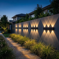 Four 6-beam solar wall wash lights mounted horizontally along long rendered boundary fence creating repeating warm diamond scalloped pattern with native grass garden path at dusk