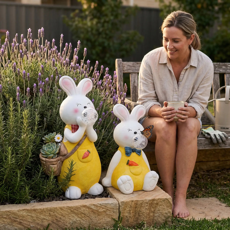 Woman sitting on a wooden garden bench smiling at OUTWAY Hoppi A004 solar bubble rabbit standing and sitting ornament pair displayed in a lavender and rosemary bed with succulents in the basket and a monarch butterfly on the sitting rabbit