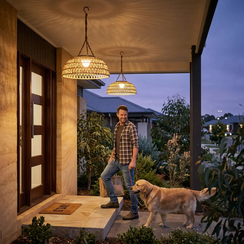 A man walking a golden retriever onto a front porch illuminated by two PT0901 Kiku Solar Rattan Lanterns, highlighting the warm welcome light and automatic dusk-to-dawn sensor.