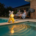 OUTWAY Hoppi A004 sitting solar bubble rabbit ornament glowing warm LED light on a pool edge at night with warm light reflecting in the water while a couple relaxes on sun loungers with wine glasses in an Australian backyard
