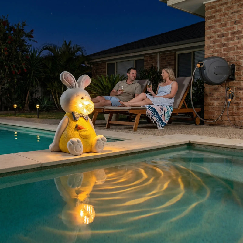 OUTWAY Hoppi A004 sitting solar bubble rabbit ornament glowing warm LED light on a pool edge at night with warm light reflecting in the water while a couple relaxes on sun loungers with wine glasses in an Australian backyard