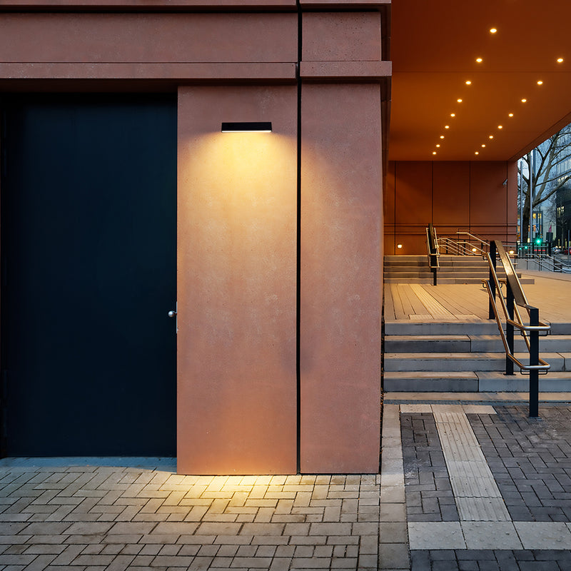 A modern rectangular wall-mounted light fixture emitting warm yellow light onto a textured terracotta-colored stucco pillar, set against an urban architectural background with steps and a covered walkway.
