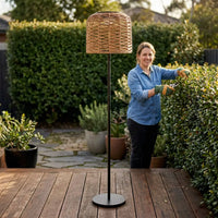 Woman gardening on a wooden deck next to a tall outdoor rattan floor lamp with a woven shade, showcasing a stylish and functional garden lighting idea.