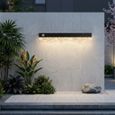 Modern outdoor wall light fixture on a concrete wall with plants and a rock in the foreground.