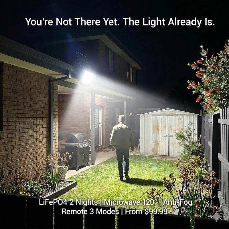 Solar flood light illuminating a backyard at night, man walking toward the house entrance, brick wall with BBQ and garden shed visible. Text: "You're Not There Yet. The Light Already Is." LiFePO4 2 Nights | Microwave 120° | Anti-Fog | Remote 3 Modes 