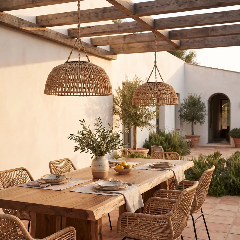 Two large woven wicker pendant lights hang over a wooden dining table set in a sunlit Mediterranean-style patio courtyard with white walls, potted plants, and a pergola.