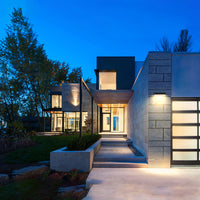 Modern single-story house exterior at dusk with stone and concrete walls, large glass windows, a lit entryway, and landscaped lawn and trees.