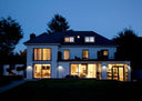 A modern two-story house at dusk with white exterior walls, large floor-to-ceiling glass windows, and warm interior lighting, set against a dark blue sky and trees.