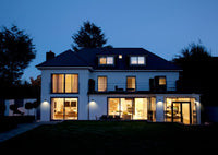 A modern two-story house at dusk with white exterior walls, large floor-to-ceiling glass windows, and warm interior lighting, set against a dark blue sky and trees.