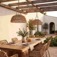 Two large woven wicker pendant lights hang over a wooden dining table set in a sunlit Mediterranean-style patio courtyard with white walls, potted plants, and a pergola.