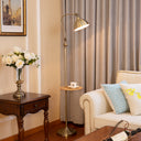 A brass floor lamp with a curved arm and adjustable shade stands in a living room beside a wooden side table with white flowers, a cream sofa with blue patterned pillows, and beige curtains in the background.
