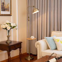 A brass floor lamp with a curved arm and adjustable shade stands in a living room beside a wooden side table with white flowers, a cream sofa with blue patterned pillows, and beige curtains in the background.